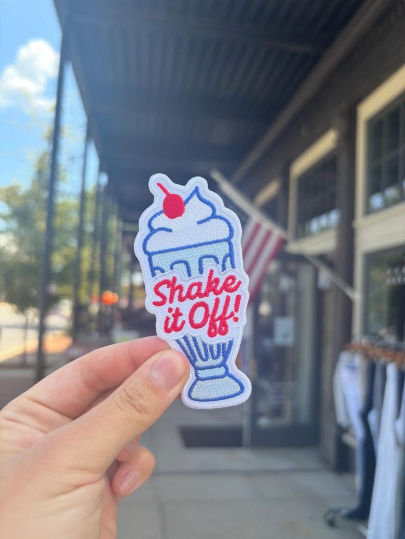 May include: Embroidered patch depicting a cartoon milkshake with a cherry. The patch has a white background with blue and red details, and the text "Shake it Off!" in red. The patch is held in front of a blurred outdoor background.