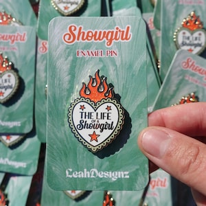 May include: Enamel pin with a heart-shaped design that reads "The Life of a Showgirl" in black text, surrounded by a gold border and topped with orange flames. The pin is on a green backing card with the word "Showgirl" in orange.