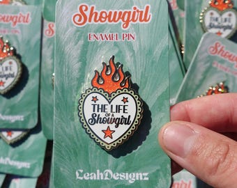 May include: Enamel pin with a heart-shaped design that reads "The Life of a Showgirl" in black text, surrounded by a gold border and topped with orange flames. The pin is on a green backing card with the word "Showgirl" in orange.