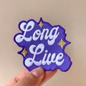 May include: A purple embroidered patch with the words "Long Live" in white script. The patch has a decorative border and gold star accents. The patch is held up by a hand.