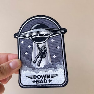 May include: Embroidered patch featuring a UFO abducting an astronaut. The patch is primarily navy blue, white, and black, with the text "DOWN + BAD" at the bottom. The design includes stars and clouds.