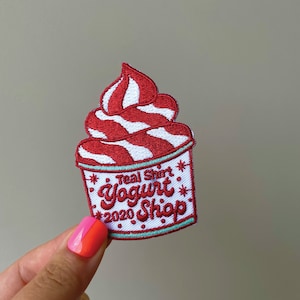Peut inclure: Écusson brodé représentant un tourbillon rouge et blanc, ressemblant à une glace à l'italienne. Le patch porte l'inscription "Teal Shirt Yogurt 2020 Shop" en script rouge sur fond blanc, avec une bordure turquoise et des étoiles.
