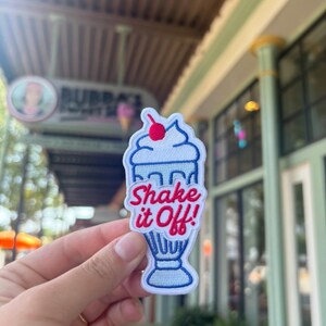 May include: Embroidered patch shaped like a milkshake, with the words "Shake it Off!" in red and blue. The patch has a cherry on top. It is held in front of a blurred outdoor background.