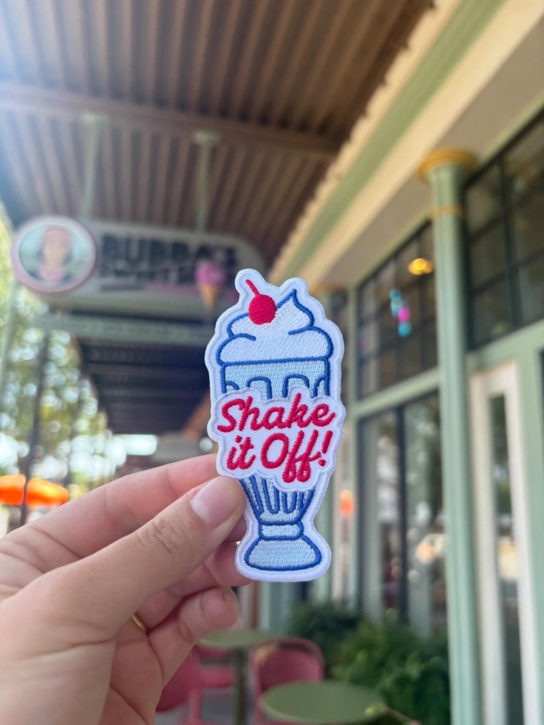 May include: Embroidered patch shaped like a milkshake, with the words "Shake it Off!" in red and blue. The patch has a cherry on top. It is held in front of a blurred outdoor background.