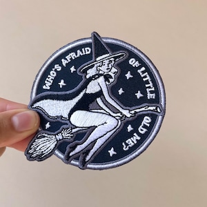 May include: Embroidered patch with a witch on a broom, the text reads "WHO'S AFRAID OF LITTLE OLD ME?". The design is in black, white, and grey, with a dark blue circular border and star accents. The witch is wearing a pointed hat.