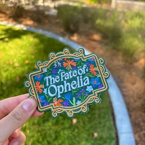 May include: Embroidered patch with the text "The Fate of: Ophelia" in white, surrounded by colorful floral designs. The patch has a green background and a decorative gold border. The patch is held against a background of green grass and foliage.