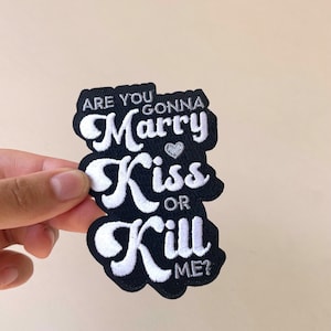 May include: Black and white embroidered patch with the text "ARE YOU GONNA Marry Kiss OR Kill ME?" in a layered design. The word "Marry" has a small heart above it. The patch is held by a hand.