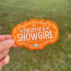 May include: An orange embroidered patch with the text "THE LIFE OF A SHOWGIRL" in white. The patch has a decorative border and star accents. The background is blurred green grass.
