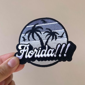 May include: Embroidered patch featuring a Florida design. The patch is circular with a black border and a stylized image of palm trees and birds against a blue and gray striped background. The word "Florida!!!" is embroidered in white.
