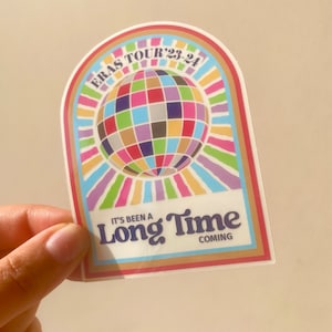 May include: A sticker with a colorful disco ball design, rainbow accents, and the text "ERAS TOUR '23-'24." The sticker also includes the phrase "It's Been a Long Time Coming."