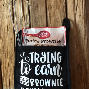 May include: Black oven mitt with the text "Trying to earn brownie points" in white. A whisk is pictured next to the text. A Betty Crocker Fudge Brownie mix package is in the pocket of the oven mitt.