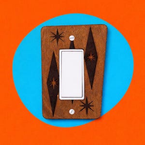 May include: A wooden light switch cover with a mid-century modern design. The cover is stained a warm brown and features etched starburst and diamond shapes. The white switch is centered, and the cover is set against a blue circle on an orange background.