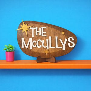 May include: Wooden sign with the text "THE McCULLYS" in white, set against a brown oval shape with starburst accents. The sign is displayed on a wooden stand, with a small potted plant in a pink pot to the left.