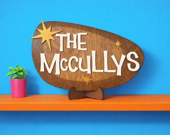 May include: A wooden sign reading "THE McCULLYS" in white, set against a brown oval shape with starburst accents. The sign is displayed on a wooden stand, with a small potted plant in a pink pot to the left.