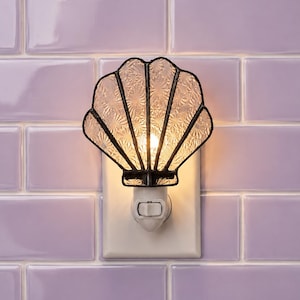 May include: A seashell-shaped night light featuring clear, textured glass panels held together by dark metal soldering. The light is plugged into a white wall outlet base with a small toggle switch, emitting a warm glow from within the shell.