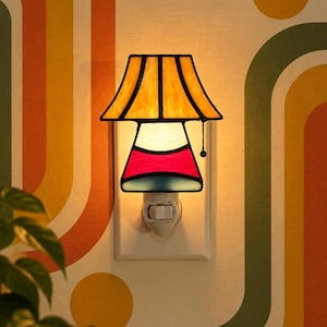 May include: A stained glass plug-in night light featuring a flared lampshade in amber yellow and a base section with red and blue glass panels. The lamp is illuminated, casting a warm glow, and includes a small black pull chain on the right side.