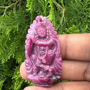 Könnte beinhalten: Ein geschnitzter, rubinroter Edelsteinanhänger, der eine Gottheitsfigur darstellt. Der Anhänger weist komplizierte Details auf, darunter eine Krone und fließende Gewänder. Der Hintergrund ist eine verschwommene Ansicht von grünem Laub.