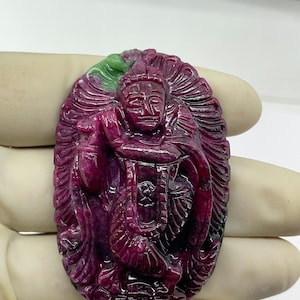 Könnte beinhalten: Ein geschnitzter Rubin-Anhänger mit einer detaillierten Darstellung einer Gottheit mit einer Krone und aufwendigen floralen Designs. Der Anhänger ist oval geformt und hat ein grün-violettes Farbschema.