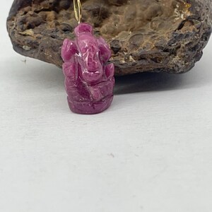 May include: A carved, magenta-colored pendant depicting the Hindu deity Ganesha. The pendant is suspended from a gold-colored loop. The background features a rough, brown stone.