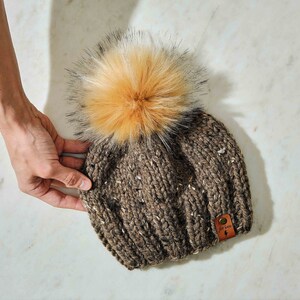 May include: A brown knit beanie with a large, fluffy, tan faux fur pom-pom. The hat has flecks of white and brown throughout the knit. A small leather tag with the text "for knits" and a lightning bolt is attached to the side.