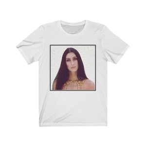 Może przedstawiać: Biały t-shirt z kwadratowym obrazem kobiety z długimi ciemnymi włosami, która nosi złotą cekinową górę na białym tle z złotym brokatem.