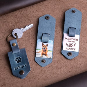 May include: A collection of personalized leather keychains in a muted blue-gray hue. One keychain features a paw print and the name "ROCKY". Another displays a photo of a dog, and the third reads "FOREVER loved ROCKY" with a paw print and wings.