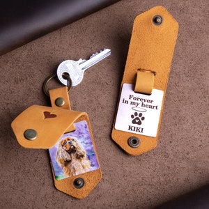 May include: Tan leather keychain with a photo of a dog inside a flap, secured with a snap. The keychain has a heart design and a metal tag that reads "Forever in my heart" with a paw print and the name "KIKI". A silver key is also present.