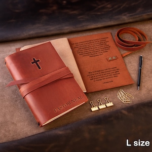 May include: A brown leather journal with a cross embossed on the front cover. The journal is closed and tied with a brown leather strap. The text "PASTOR NEAL" is engraved on the front cover. The journal is sitting on a brown surface with a pen, paper clips, and a leather strap.