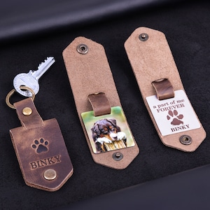 May include: Leather keychains with a dog theme. One keychain has a paw print and the name "BINKY" engraved. Another keychain features a color photo of a dog. The third keychain has the text "a part of me FOREVER" and the name "BINKY".