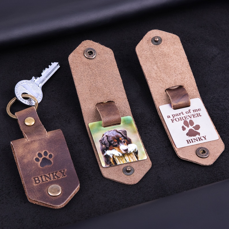 May include: Leather keychains with a dog theme. One keychain has a paw print and the name "BINKY" engraved. Another keychain features a color photo of a dog. The third keychain has the text "a part of me FOREVER" and the name "BINKY".