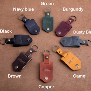 May include: A collection of leather keychains in various colors, including black, navy blue, green, burgundy, dusty blue, brown, copper, and camel. Each keychain is engraved with the word "DADDY" and features a metal ring and snap closure.
