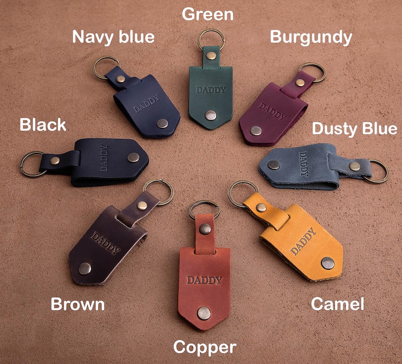 May include: A collection of leather keychains in various colors, including black, navy blue, green, burgundy, dusty blue, brown, copper, and camel. Each keychain is engraved with the word "DADDY" and features a metal ring and snap closure.