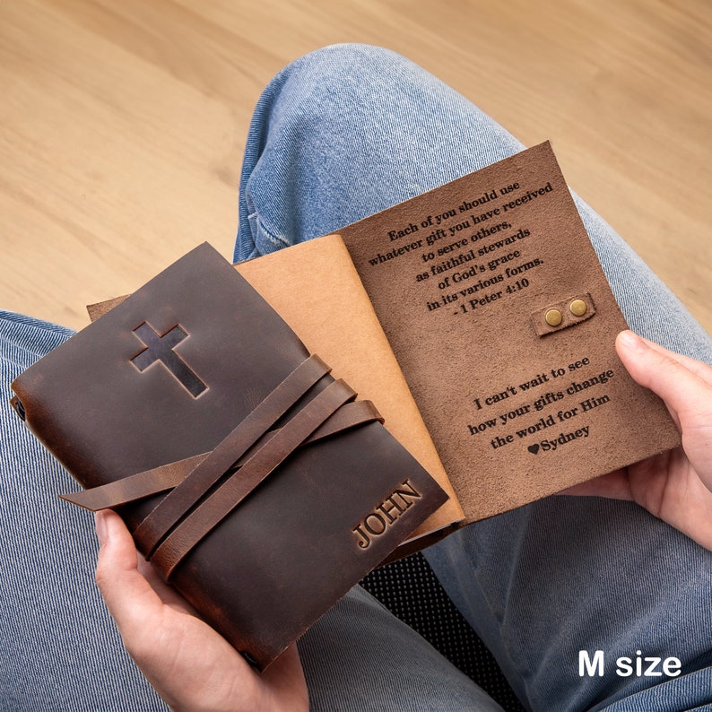 May include: A brown leather journal with a cross embossed on the front cover. The journal is closed and has a leather strap with the name "JOHN" embossed on it. The journal is open to reveal a brown leather interior with a quote from the Bible, "Each of you should use whatever gift you have received to serve others, as faithful stewards of God's grace in its various forms. -1 Peter 4:10". The journal also has a handwritten message that says "I can't wait to see how your gifts change the world for Him. Sydney".