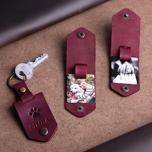 May include: Burgundy leather keychains and photo holders. One keychain has a paw print and the name "SIMBA". Two photo holders display color and black-and-white photos. All pieces have metal snap closures and rivets.