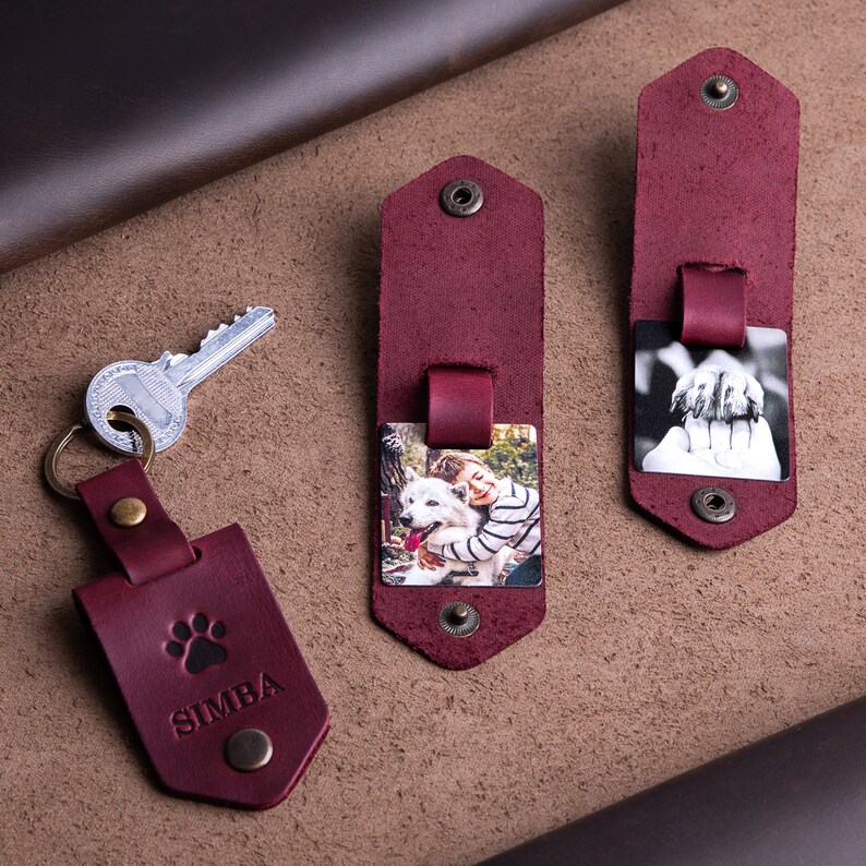 May include: Burgundy leather keychains and photo holders. One keychain has a paw print and the name "SIMBA". Two photo holders display color and black-and-white photos. All pieces have metal snap closures and rivets.
