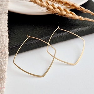 Peut inclure: Une paire de boucles d'oreilles créoles géométriques dorées de forme carrée. Les boucles d'oreilles sont sur une surface blanche.