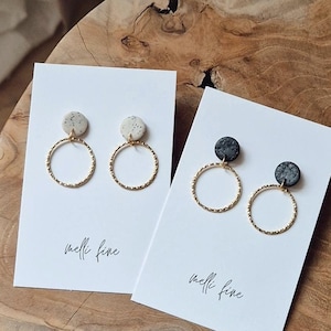 Könnte beinhalten: Zwei Paar Ohrringe auf weißen Karten. Die Ohrringe sind goldene Creolen mit kleinen runden weißen und schwarzen, gesprenkelten Keramik-Scheiben. Die Karten tragen den Text "melli fine" aufgedruckt.