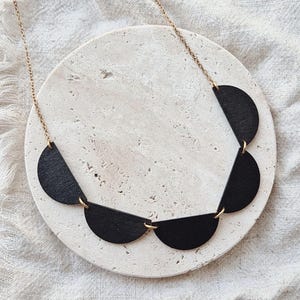 Puede incluir: Un collar de madera negra con una cadena dorada. El collar presenta una serie de colgantes de madera semicirculares conectados por pequeños anillos dorados. El collar se exhibe sobre una piedra blanca y circular.