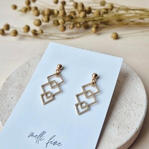 Könnte beinhalten: Goldfarbene geometrische Ohrringe mit quadratischem Design. Die Ohrringe werden auf einer weißen Karte mit dem handgeschriebenen Text "melli fine" präsentiert. Der Hintergrund umfasst getrocknete Pflanzen und eine neutralfarbene Oberfläche.