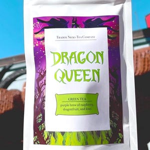 May include: A white pouch of Dragon Queen green tea. The label features a purple and green design with the text "DRAGON QUEEN" in large, stylized letters. The label also lists the tea's ingredients: raspberry, dragonfruit, and kiwi.