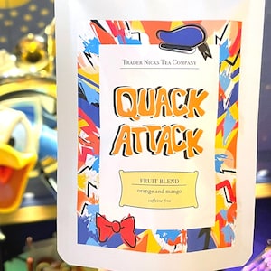 May include: White tea bag with the text "QUACK ATTACK" in orange and black. The bag also says "FRUIT BLEND orange and mango caffeine-free". The bag has a colorful design with a blue hat and red bow.