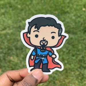 May include: A sticker featuring a cartoon illustration of Doctor Strange. The sticker shows Doctor Strange in a blue and black outfit with a red cape. The sticker is held in a hand against a green background.