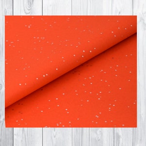 May include: Vibrant orange fabric with a scattering of tiny white dots. The material is folded, revealing its texture and form. The background features a light wood grain pattern.