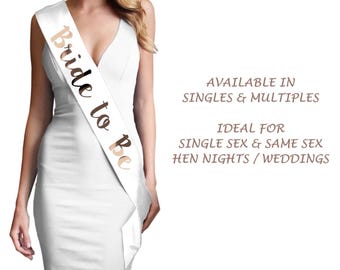 May include: A white satin sash with the words "Bride to Be" in a copper-colored script. The sash is draped over a white dress. The text "Available in Singles & Multiples" is also visible.