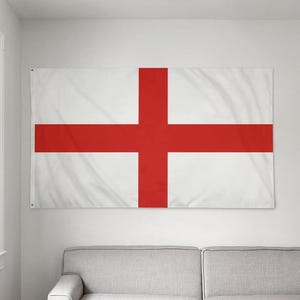 May include: A rectangular white flag featuring a centered red cross, the flag of England, hangs on a wall. The flag is a simple design, with the red cross prominent against the white background. The flag is displayed indoors.