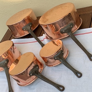 May include: Five copper saucepans with dark metal handles. The pans have a shiny surface and are arranged on a white cloth with a red stripe. The handles are riveted to the copper bodies. A set of kitchenware.