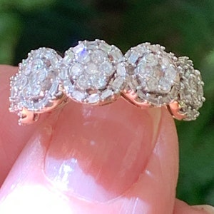 May include: A gold ring with a series of diamond clusters. Each cluster is made up of round diamonds, surrounded by smaller, rectangular diamonds. The ring is held up against a blurred green background. The ring is a piece of jewellery.