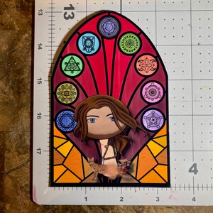 Puede incluir: Una pegatina de vitral con una mujer de cabello largo y casta&ntilde;o y un gato. La mujer lleva un atuendo de cuero marr&oacute;n y el gato es blanco con manchas marrones. El dise&ntilde;o del vitral presenta un fondo rojo con formas geom&eacute;tricas de colores.