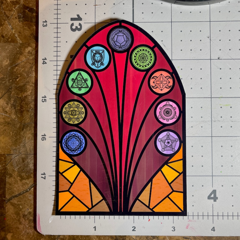 Puede incluir: Una pegatina de ventana de vitral con fondo rojo y un dise&ntilde;o colorido de formas geom&eacute;tricas y s&iacute;mbolos. La pegatina tiene forma de arco y un contorno negro.