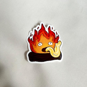 Op de afbeelding: Een sticker met een cartoonvlamkarakter met een verraste uitdrukking. De vlam is voornamelijk rood en oranje, met een gele tongachtige verlenging. Het karakter zit op een donkerbruin blok hout.